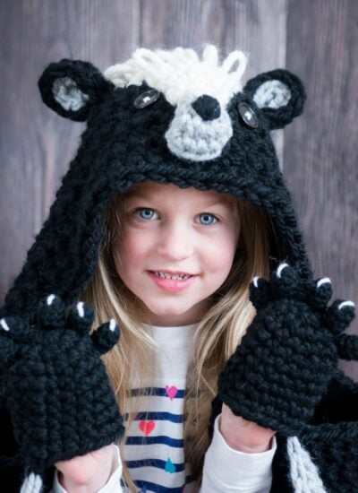 A little girl wearing a crocheted black bear hoodie, adorned with a hooded woodland skunk blanket.