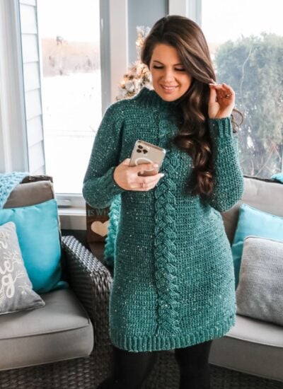 A woman wearing a teal cable knit sweater, demonstrating how to make The Piece of Cake Cardi in front of a fireplace.