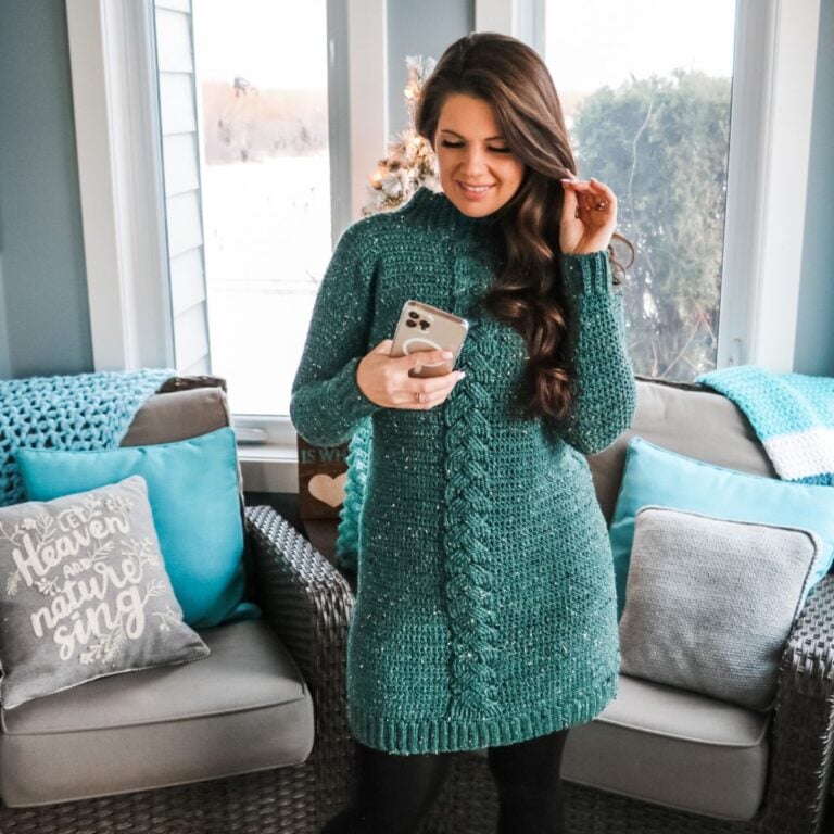 A woman wearing a teal cable knit sweater, demonstrating how to make The Piece of Cake Cardi in front of a fireplace.