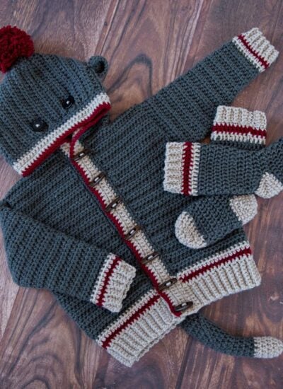 A knitted sock monkey sweater and mittens on a wooden floor, showcasing the Step-By-Step Guide to Make The Piece of Cake Cardi.