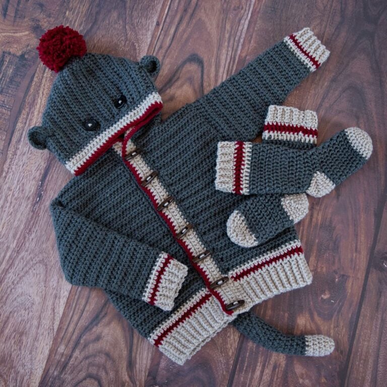 A knitted sock monkey sweater and mittens on a wooden floor, showcasing the Step-By-Step Guide to Make The Piece of Cake Cardi.