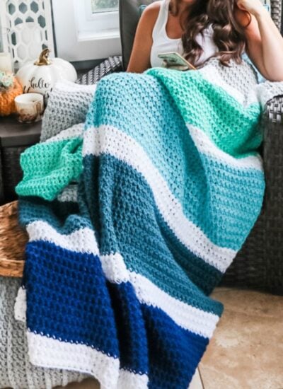 A woman sitting in a chair with a crocheted blanket, working on her handmade "Piece of Cake Cardi.