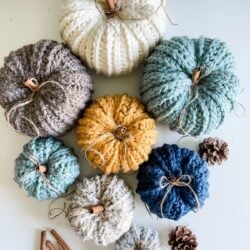 Seven rustic crocheted pumpkins in various colors, with cinnamon sticks as stems, are arranged on a table alongside pine cones and extra cinnamon sticks.