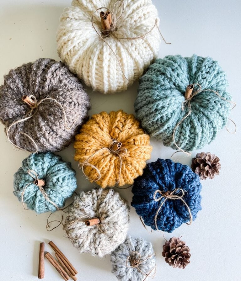 Seven rustic crocheted pumpkins in various colors, with cinnamon sticks as stems, are arranged on a table alongside pine cones and extra cinnamon sticks.