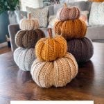 Three stacks of Crochet Rustic Farmhouse Pumpkins in neutral and orange tones sit on a wooden table in a cozy living room with a sofa, pillows, and a cow painting in the background.
