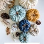 Assorted Crochet Rustic Farmhouse Pumpkins in various colors and sizes are arranged on a table with pinecones, cinnamon sticks, wooden beads, and dried flowers.