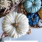 A hand holds a chunky, cream-colored crocheted pumpkin with a cinnamon stick stem, surrounded by rustic Crochet Farmhouse Pumpkins and dried flowers in woven baskets.