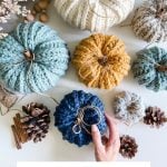 Assorted Crochet Rustic Farmhouse Pumpkins in various colors are arranged with pinecones, wooden beads, and dried flowers on a white surface. A hand holds a dark blue pumpkin. Text reads "Rustic Farmhouse Pumpkins.
