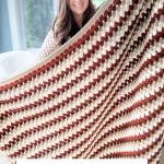 A woman holds up a large crocheted granny blanket in fall colors with the text "FALL FOR GRANNY BLANKET Free Crochet Pattern & Video" displayed, perfect to pair with Crochet Rustic Farmhouse Pumpkins for cozy autumn decor.