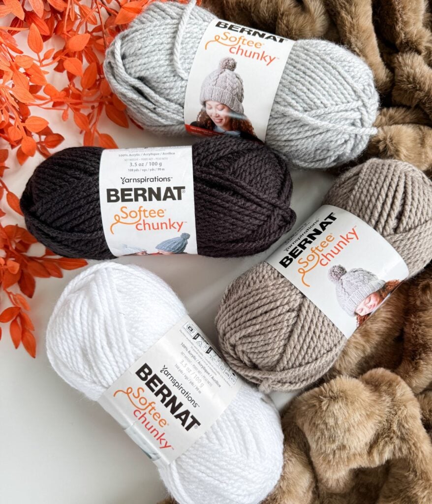 Four skeins of Bernat Softee Chunky yarn in white, taupe, gray, and dark brown are arranged on a faux fur surface next to orange artificial leaves, perfect for projects or as inspiration for your next yarn substitution guide.
