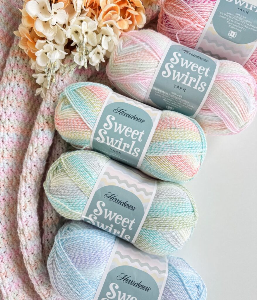 Three skeins of Herschners Sweet Swirls pastel yarn and one pink yarn rest on a crocheted blanket near artificial flowers—a cozy scene perfect for exploring your next project with a handy yarn substitution guide.