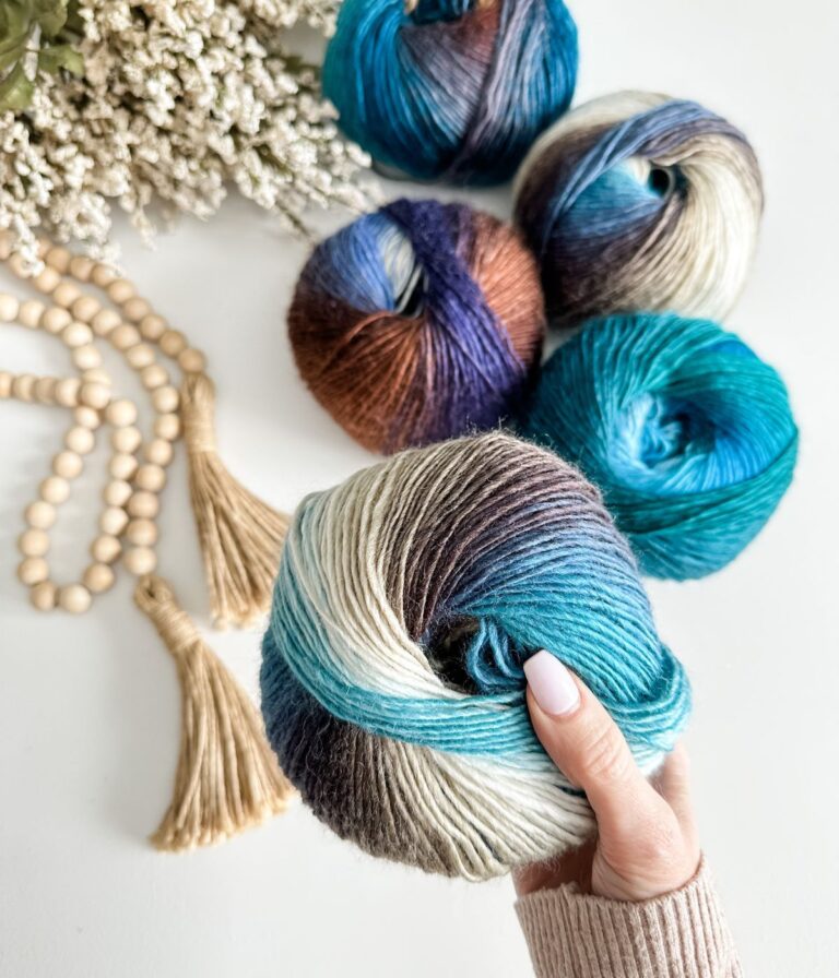 A hand holds a skein of multicolored yarn, with four similar skeins, wooden beads, tassels, and white flowers arranged on a white surface—a perfect setup for exploring a yarn substitution guide.