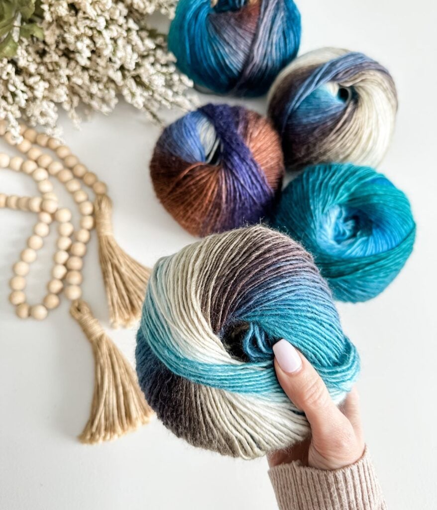 A hand holds a skein of multicolored yarn, with four similar skeins, wooden beads, tassels, and white flowers arranged on a white surface—a perfect setup for exploring a yarn substitution guide.