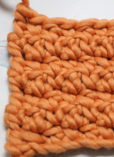 Close-up of an orange, chunky knit swatch with a textured pattern, shown on a white surface—perfect inspiration if you want to learn how to crochet the seed stitch.