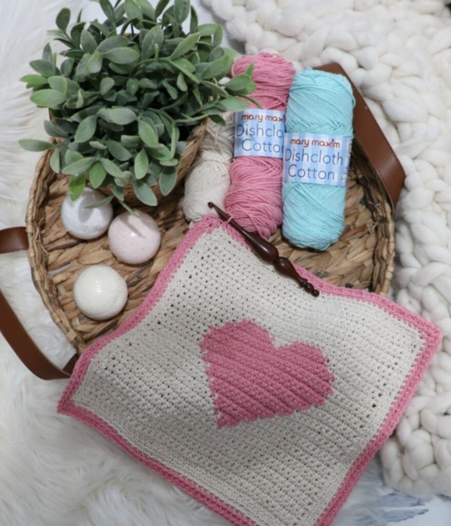 A woven tray holds three yarn balls, a potted green plant, round bath bombs, and a handmade crocheted square with a pink heart and border—perfect inspiration for your next Crochet Heart Dishcloth Pattern.