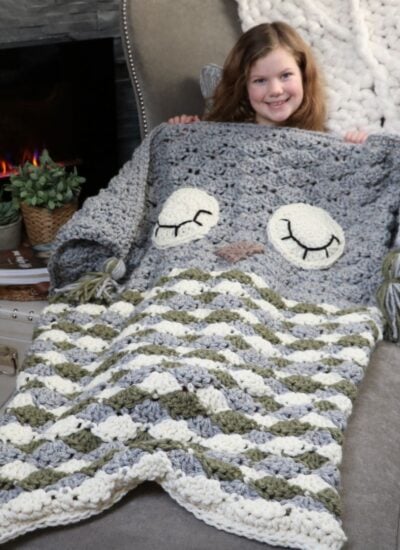 A sleepy little girl snuggles up with her crocheted owl cocoon blanket in front of a cozy fireplace.
