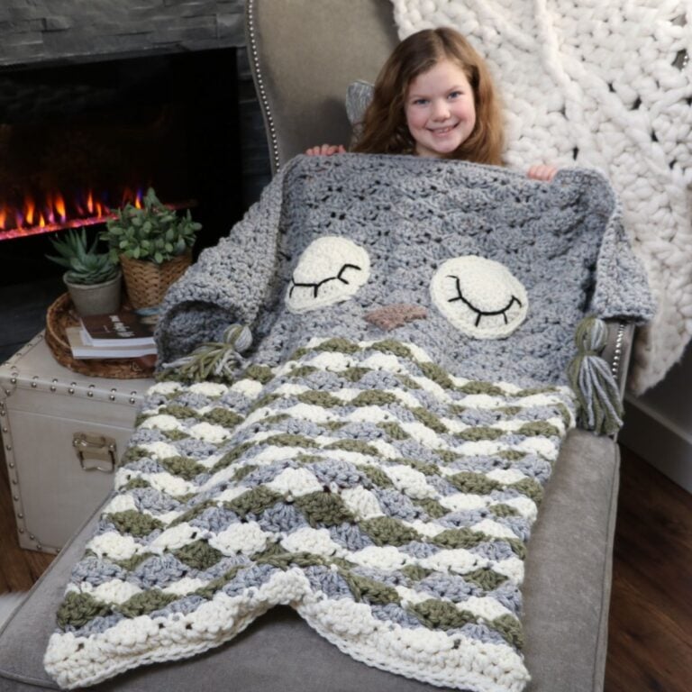 A sleepy little girl snuggles up with her crocheted owl cocoon blanket in front of a cozy fireplace.