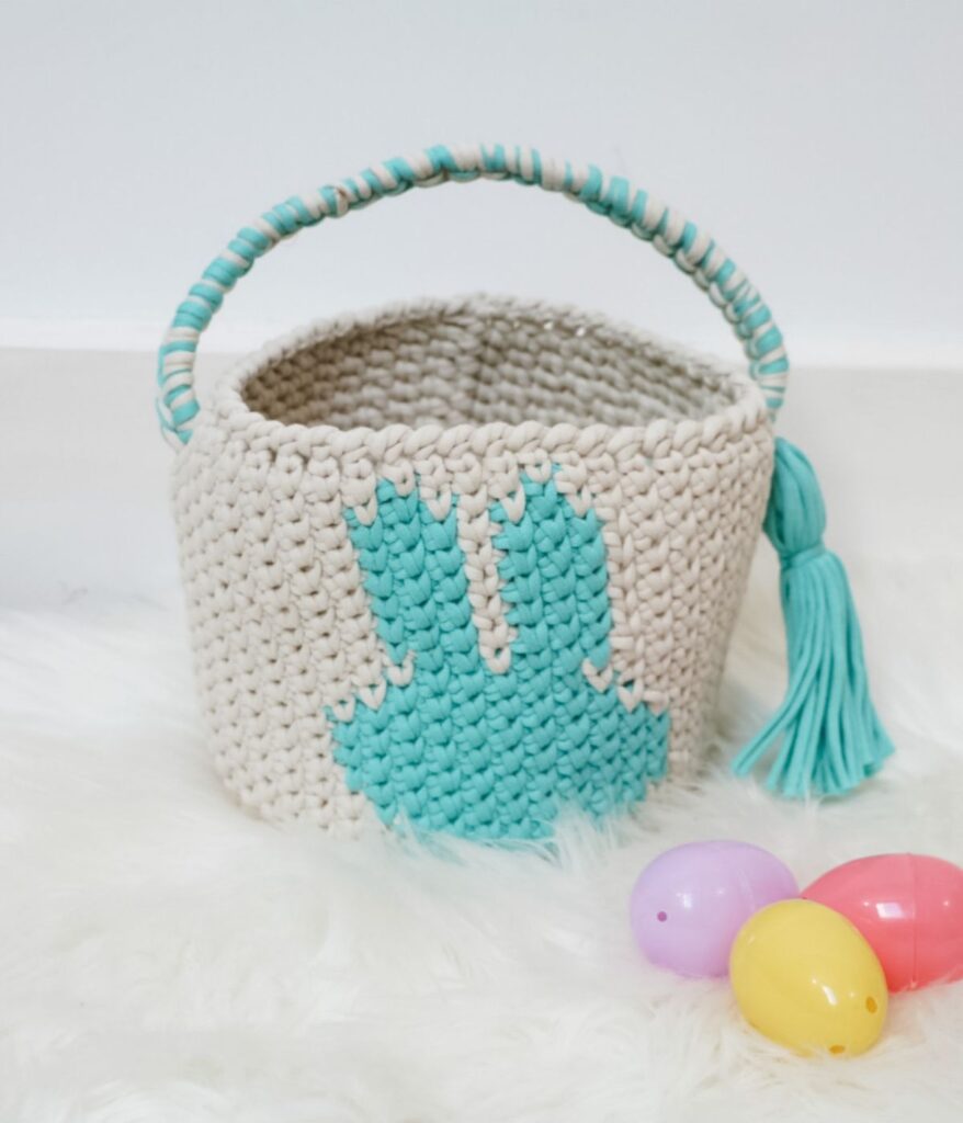 A crocheted Easter Bunny Basket with a blue bunny design, handle, and tassel sits on a white surface beside three plastic Easter eggs in purple, pink, and yellow.