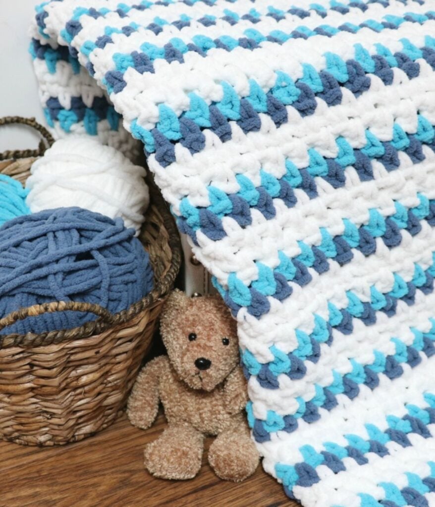 The Seaside Cuddles Baby Blanket in white, blue, and teal stripes drapes over a basket of yarn, with a small brown teddy bear sitting beside it on a wooden floor.