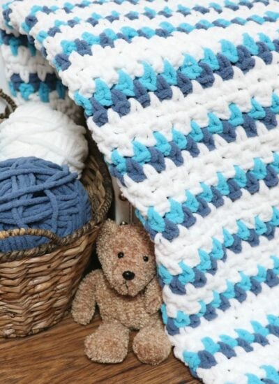 The Seaside Cuddles Baby Blanket, in delicate shades of blue and white, features a charming teddy bear nestled within a cozy basket.