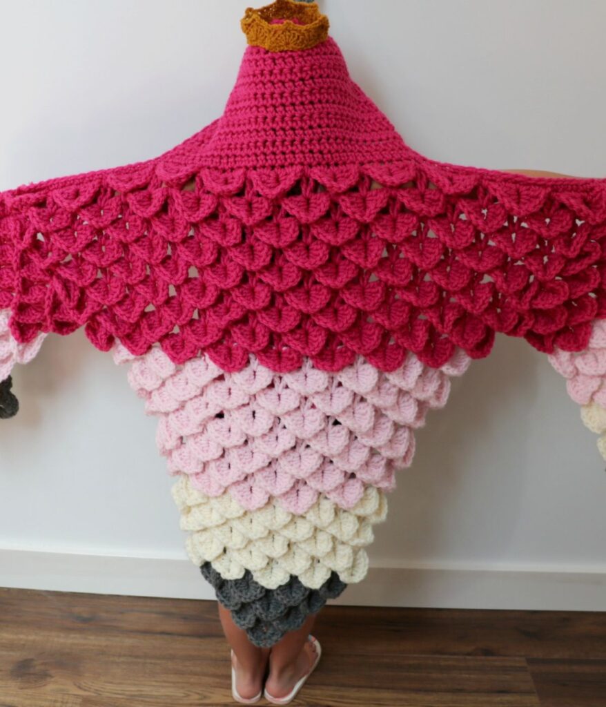 Person wearing a large, crocheted garment resembling an ice cream cone with scalloped pink, white, and brown sections—styled like a whimsical Hooded Flamingo Blanket Costume—standing against a plain wall.