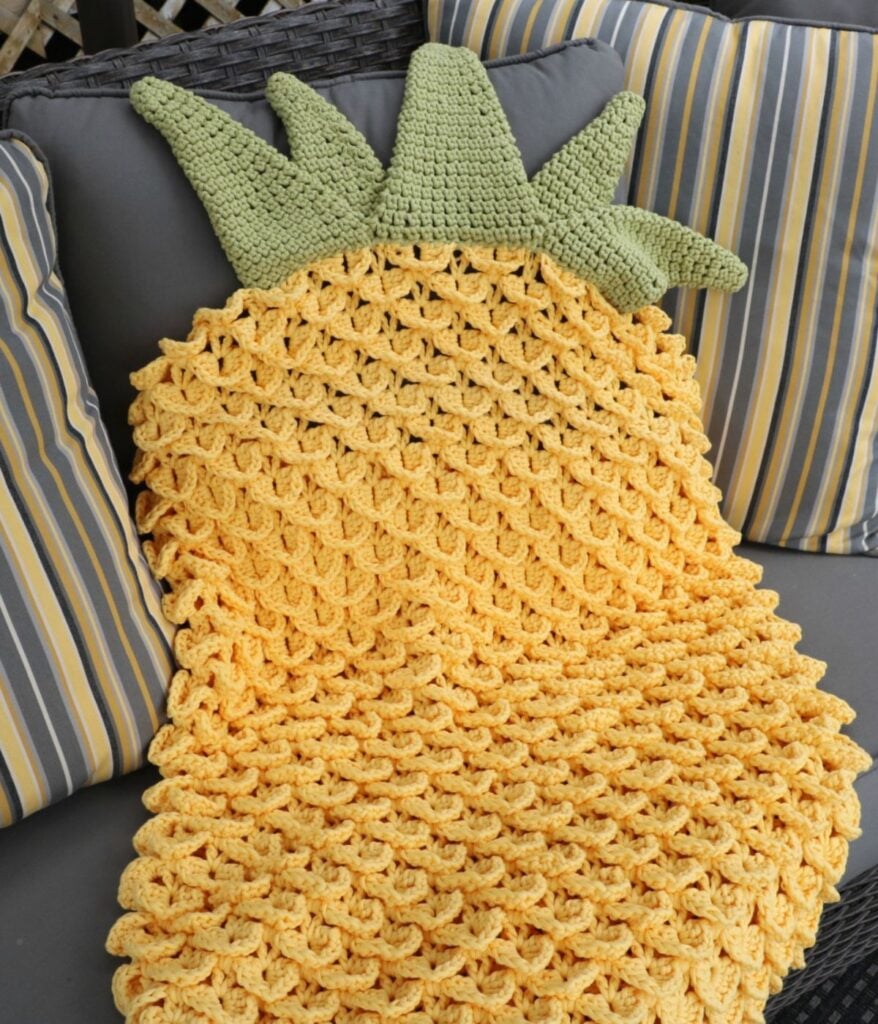A Crochet Pineapple Blanket Cocoon, featuring a textured yellow body and green leaves, is draped over a gray outdoor sofa with striped cushions.