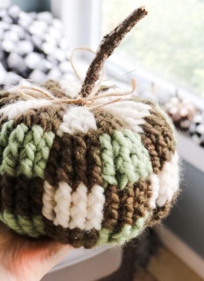 A bulky crocheted pumpkin is being held up in front of a window.