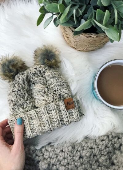 A person holding a Hooded Lion Cowl beanie and a cup of coffee.