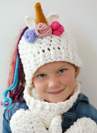 A little girl wearing a Hooded Lion Cowl and mittens adorned with a crocheted unicorn design.