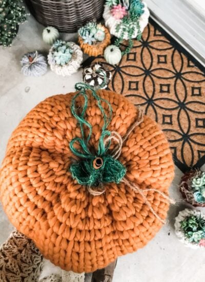 A person is holding a rustic knit pumpkin on a porch.