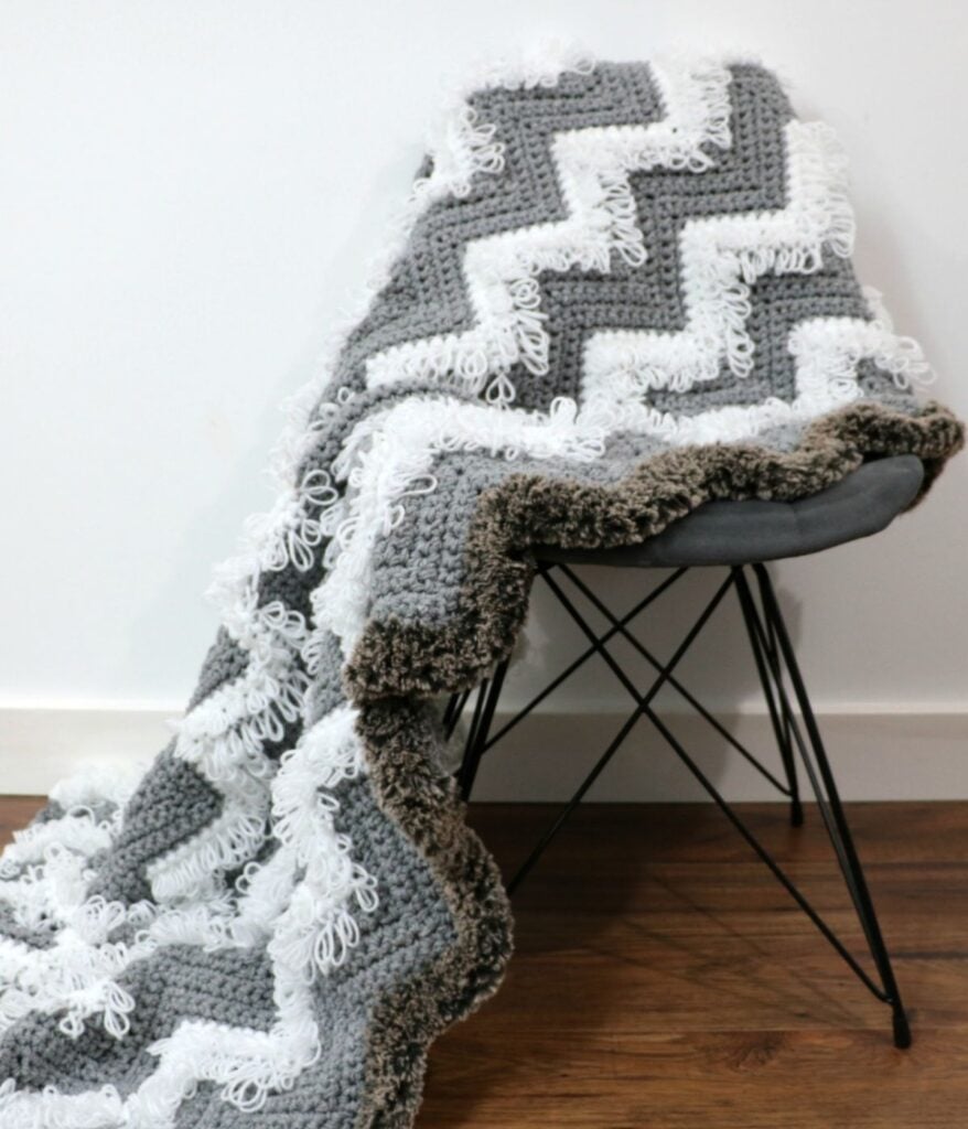 A gray and white Loopy Chevron Stitch Blanket with a zigzag pattern is draped over a dark gray chair on a wooden floor against a white wall.