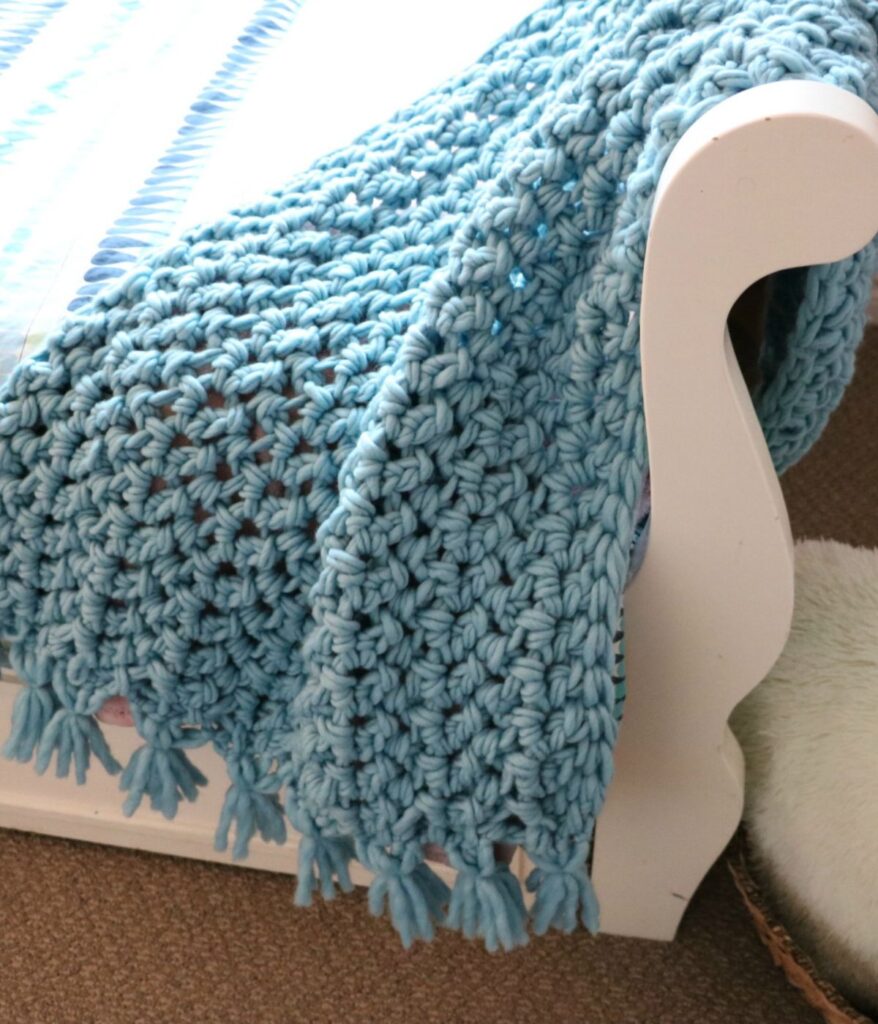 A chunky light blue Super Bulky Herringbone Throw with tassels draped over the arm of a white wooden chair or bed.
