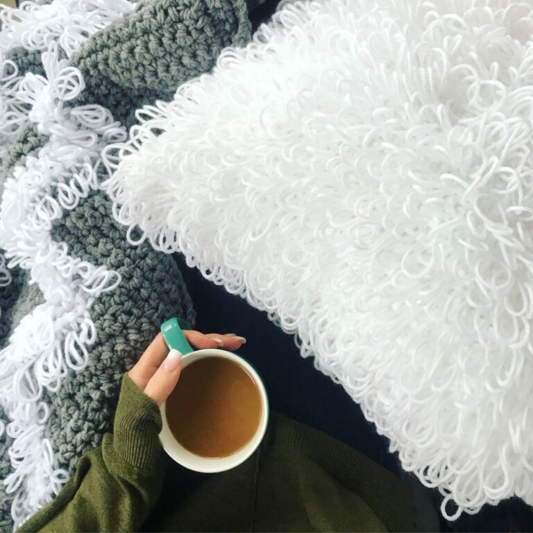 A person holding a cup of coffee beside a crocheted Loopy Stitch Pillow Cover.