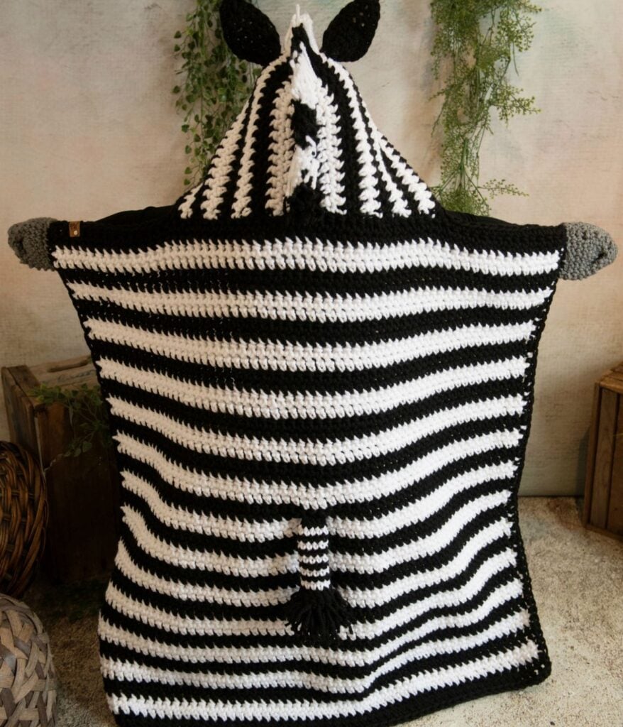 A person is wrapped in a cozy hooded zebra blanket, crocheted with black and white stripes, a tail, and a hood featuring playful zebra ears.