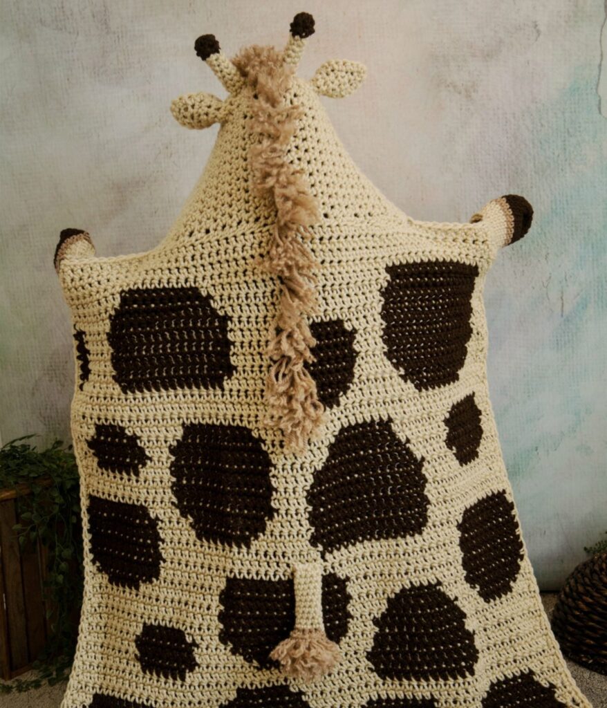 A Hooded Giraffe Blanket, crocheted to resemble a giraffe’s back, features brown spots, small ears, and a cozy yarn mane and tail for extra charm.