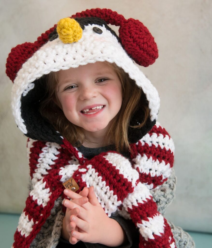 Young child smiling, wrapped in a Crochet Penguin Blanket designed as a red, white, and black hooded cape with a penguin face and earmuffs.