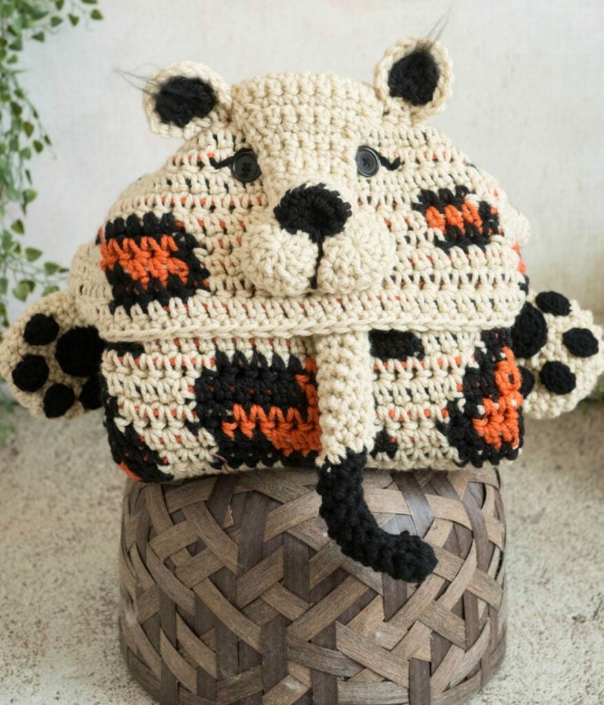 A crocheted pouch designed to resemble a leopard's face and paws sits on top of a woven basket, making a charming pair with a cozy hooded Leopard Blanket.