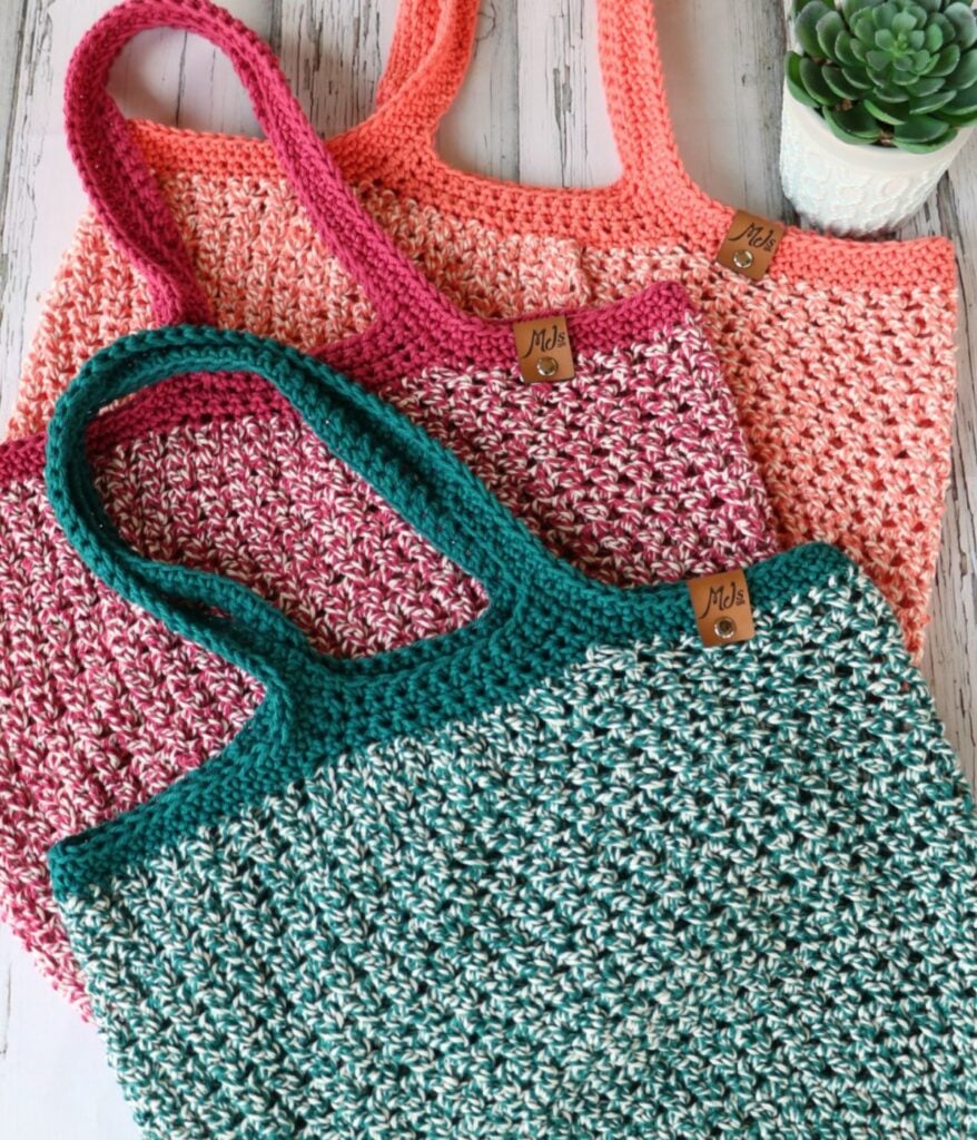 Three Dishie-lous Market Bags in coral, pink, and teal are stacked on a white wood surface near a small potted succulent.