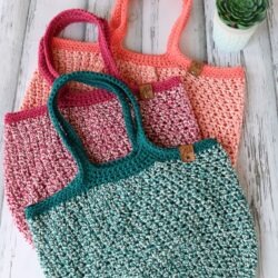 Three Crochet Market Bags in teal, pink, and coral are stacked on a white wooden surface next to a small potted succulent.