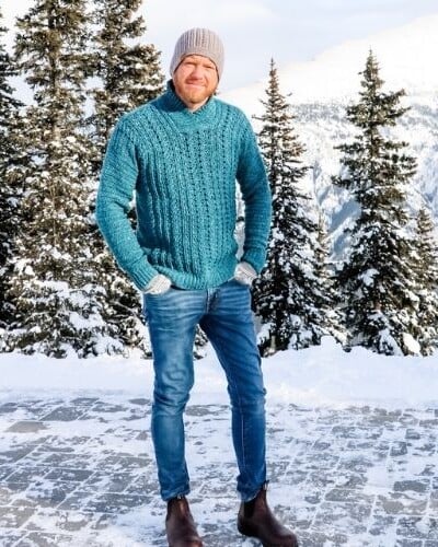 A dapper man in a blue sweater standing in front of a snow covered mountain.