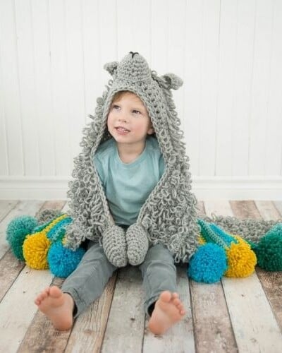 A little boy sitting on the floor with a crocheted owl hoodie and a hooded blanket.