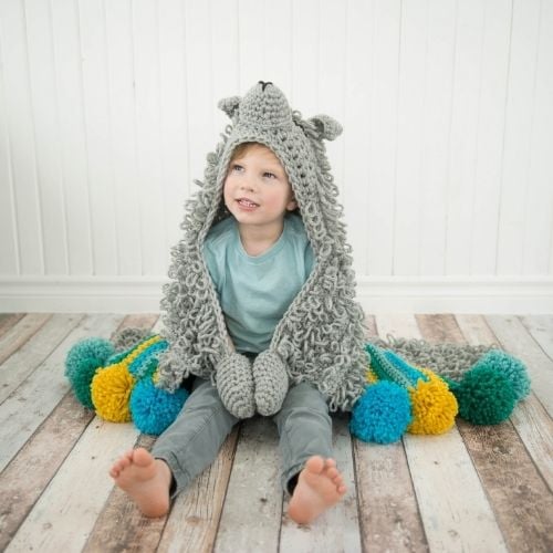 A little boy sitting on the floor with a crocheted owl hoodie and a hooded blanket.