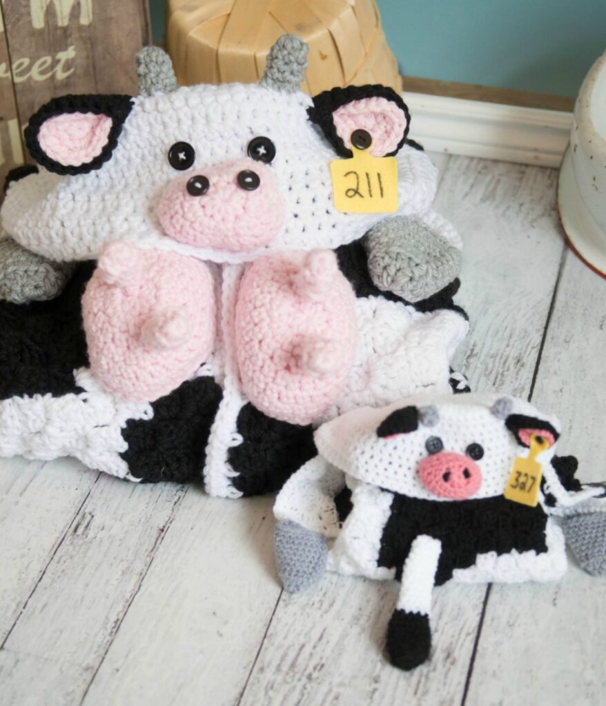 Two crocheted cow plushies, one large and one small, both with pink snouts, gray hooves, and yellow ear tags labeled "211" and "327," are displayed on a wooden surface beside a cozy Hooded Cow Blanket.