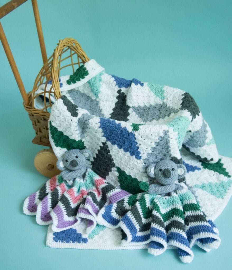 A Modern Herringbone Baby Blanket with a geometric pattern in white, gray, blue, and green is draped over a basket. Two crocheted koala loveys rest atop the blanket.