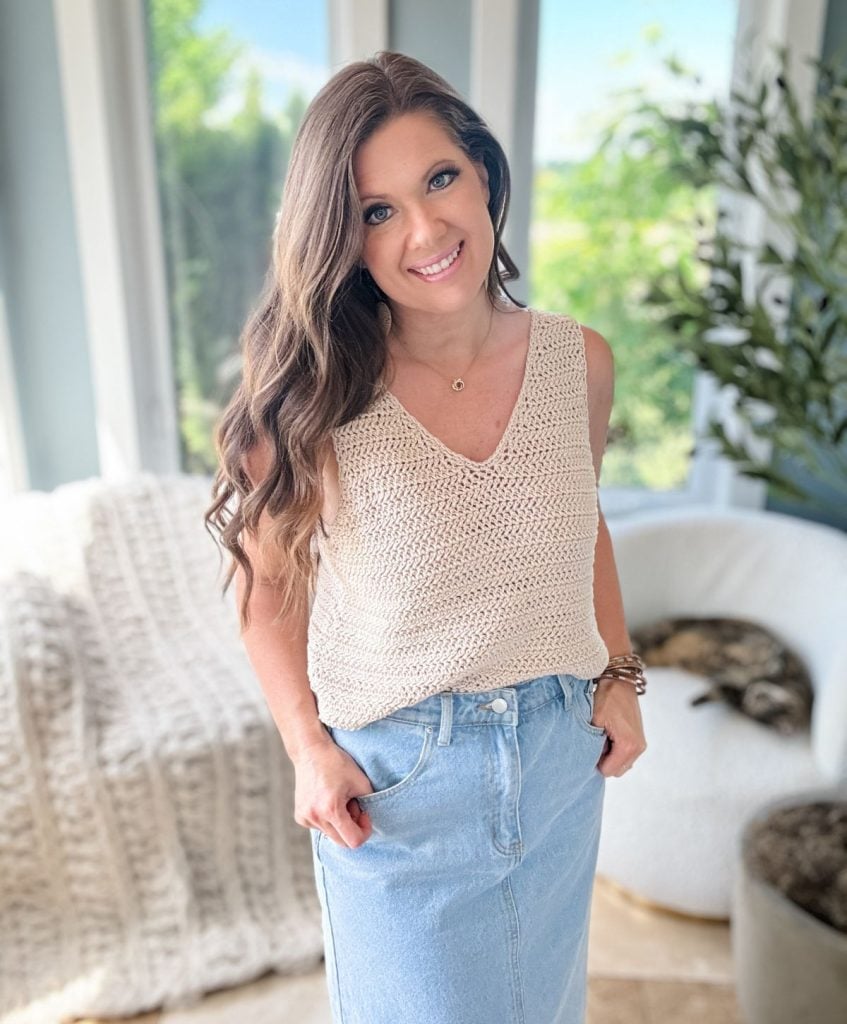 A woman with long hair wearing a crocheted Ruby-V Tank top and denim skirt stands indoors, smiling at the camera. A chair with a knitted blanket and a window view of greenery are in the background.