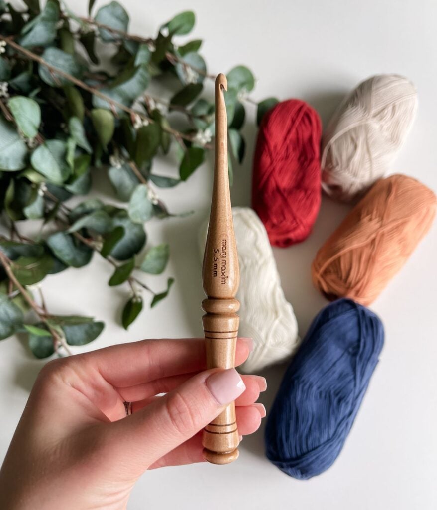 Hand holding a wooden crochet hook with balls of yarn in red, white, orange, and blue beside a branch with green leaves in the background—perfect for creating projects like the Crochet the Ruby-V Tank.