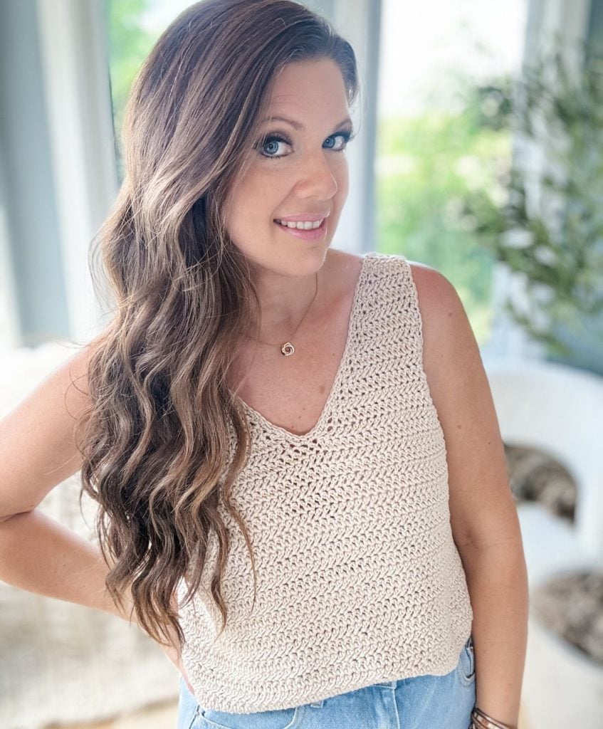 Person with long wavy hair wearing a sleeveless knit top and jeans, standing indoors with one hand on hip, smiling at the camera, as if ready to Crochet the Ruby-V Tank.