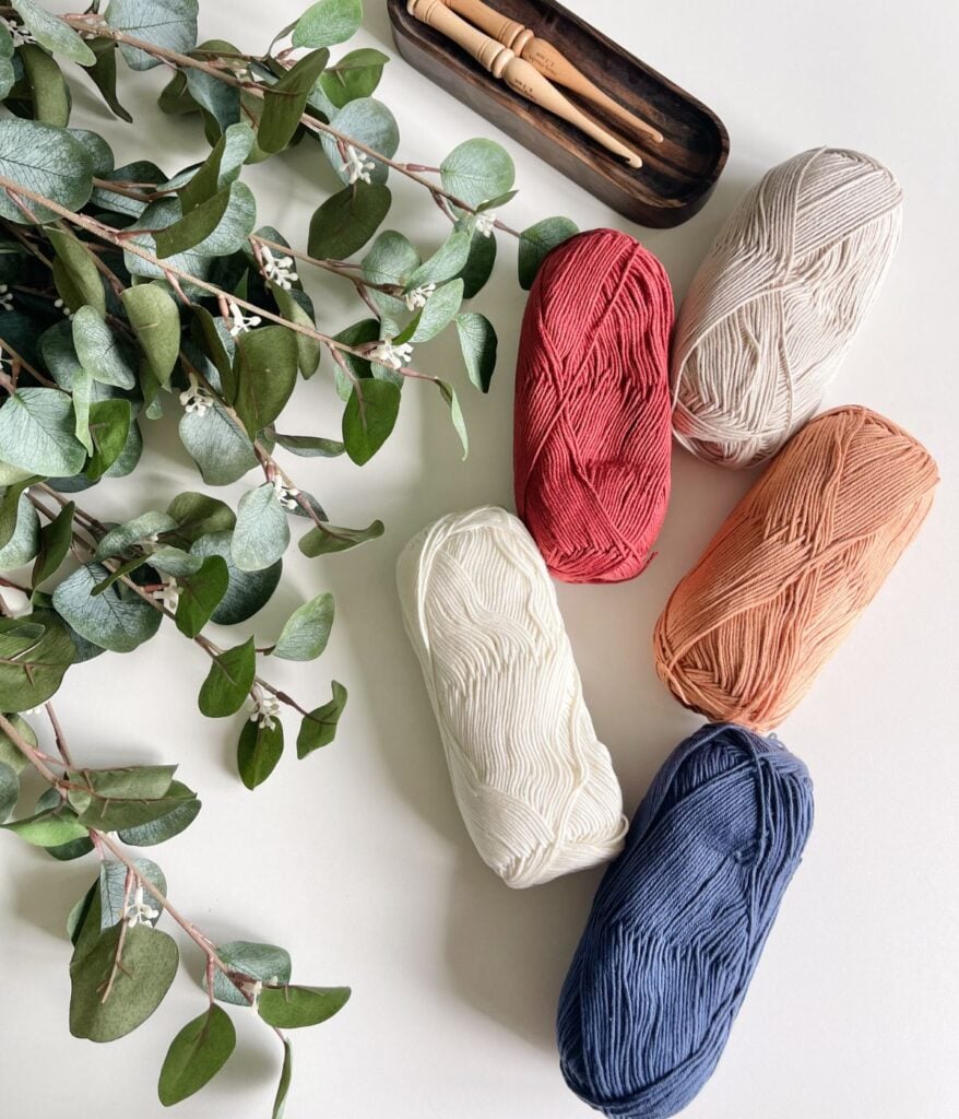 Five skeins of yarn in various colors and two wooden crochet hooks are arranged on a white surface, ready to help you Crochet the Ruby-V Tank. A sprig of eucalyptus lies to the side, adding a touch of nature.