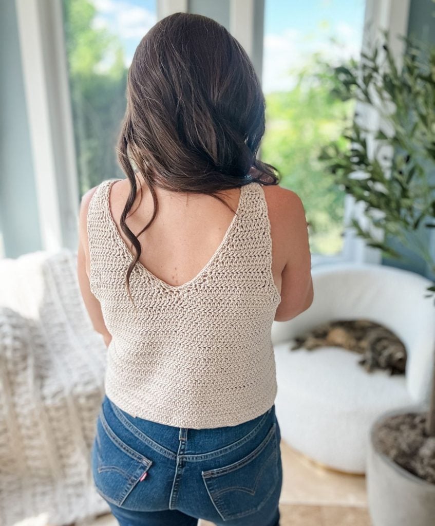 With her long hair cascading down her back, she gazes out the window in a sleeveless knitted top. Near her, a cat lounges on a chair amidst lush greenery—a scene reminiscent of the relaxed elegance of the Crochet the Ruby-V Tank pattern.