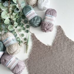 A partially completed Crochet the Ruby-V Tank lies next to several skeins of yarn in various colors, a crochet hook, and a sprig of greenery on a white surface.
